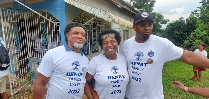 alt text: Three generations of the Henry family posing outdoors in Jamaica, wearing personalized family reunion t-shirts for a family link-up event.