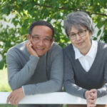 An elderly Japanese couple is smiling at the camera. They both wear glasses and a grey sweaters. They are in a garden, green tree branches behind them. They are leaning on a white barrier.