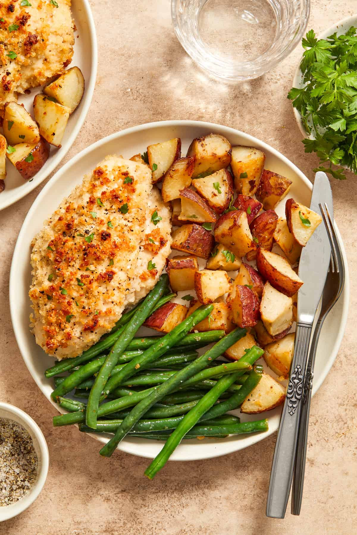 Parmesan baked chicken, roasted potatoes and green beans on white plate with fork and knife.