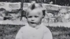 Susan as a baby wearing a white jumper and sitting on grass