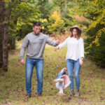 A father looking exasperated while holding a crying baby, with the mother laughing in the background. The alt text is: Exasperated father holding a crying baby during family photo shoot, with the mother laughing, capturing a funny family moment