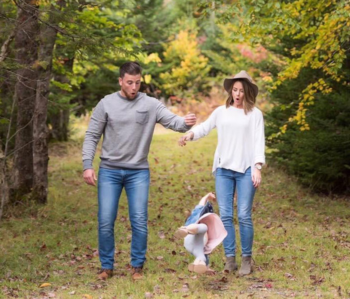 A father looking exasperated while holding a crying baby, with the mother laughing in the background. The alt text is: Exasperated father holding a crying baby during family photo shoot, with the mother laughing, capturing a funny family moment
